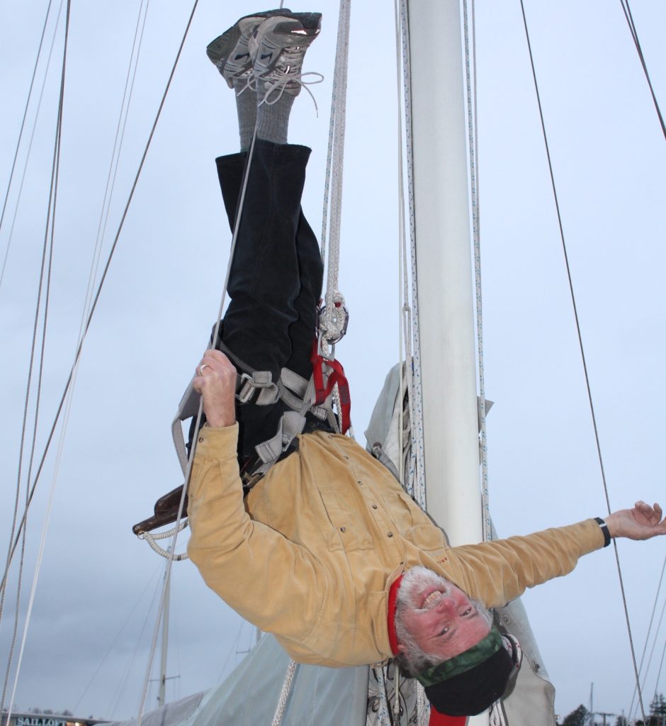 Brion Toss hanging upside down in a harness from a sailboat mast.